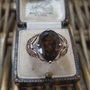 May include: A silver ring featuring a large, oval, dark brown gemstone. The ring has an intricate, swirling pattern around the stone. It is presented in a vintage, dark blue jewellery box with a cream interior.