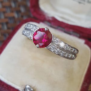 Peut inclure: Une bague argentée ornée d'une grande pierre précieuse rouge foncé, ronde et à facettes, fixée au centre. L'anneau est divisé en deux rangées parallèles, chacune incrustée de multiples petites pierres claires et scintillantes sur les côtés.