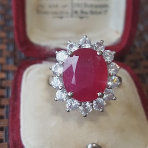 May include: A silver-toned ring featuring a large, oval-cut, deep red center stone surrounded by a halo of twelve small, round, clear, faceted stones, presented in a red velvet-lined jewelry box.