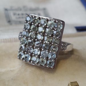 May include: A silver-toned ring featuring a large, rectangular face densely set with numerous round, pale green faceted stones arranged in a grid pattern. The band is smooth and metallic, supporting the prominent, multi-stone rectangular head.