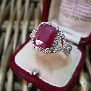May include: A silver ring featuring a large, red, square-shaped ruby surrounded by a halo of smaller diamonds. The ring is set in a white metal band with a twisted design.