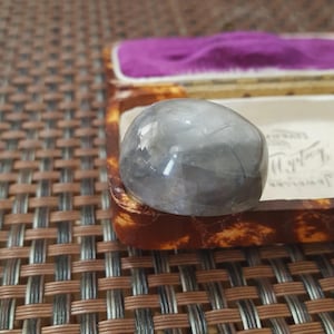 May include: A grey, rounded gemstone rests in a vintage jewellery box with a purple velvet interior. The box has a tortoiseshell pattern and a white label with cursive text. The gemstone is smooth and translucent, reflecting light.