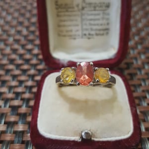 May include: A silver ring featuring three gemstones. The central, oval-cut stone is peach-coloured, with two yellow, round gemstones either side. The ring is presented in a red velvet box.