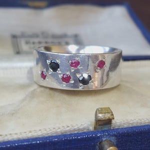 Puede incluir: Un anillo de banda ancha de plata con una disposición dispersa de piedras preciosas engastadas. El diseño incluye tres rubíes rojos redondos y dos piedras de ónice negro redondas, cada una asegurada en un engaste de estrella de cuatro puntas.