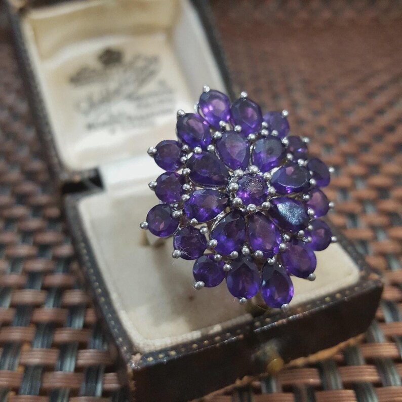 K&ouml;nnte beinhalten: Ein Silberring mit einem floralen Muster, besetzt mit zahlreichen oval geschliffenen, tiefvioletten Amethyst-Edelsteinen. Der Ring wird in einer Vintage-Schmuckschatulle aus braunem Leder pr&auml;sentiert.