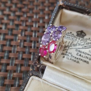 May include: Two silver rings with oval-cut gemstones. One ring features purple gemstones, the other has red gemstones. The rings are displayed inside a vintage jewelry box with printed text.