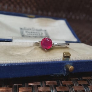 May include: A silver ring featuring a vivid red gemstone, presented in a blue jewellery box with a light-coloured interior. The ring's band has a flowing design, accentuating the central stone. The box is secured with a gold-toned clasp.