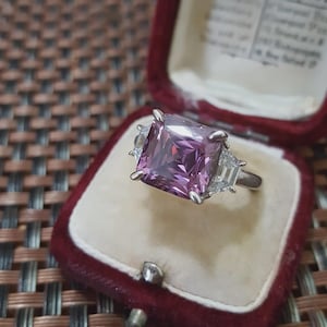 May include: A silver ring with a large, square, light purple gemstone and two smaller, white, rectangular gemstones on either side. The ring is displayed in a red velvet ring box.