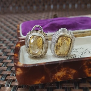 Könnte beinhalten: Ein Paar silberfarbene Ohrringe mit großen, rechteckigen, gelben Edelsteinen. Die Ohrringe sind in einem silbernen Rahmen mit einem strukturierten Rand gefasst. Präsentiert in einer Vintage-Schmuckschatulle mit Schildpattmuster.