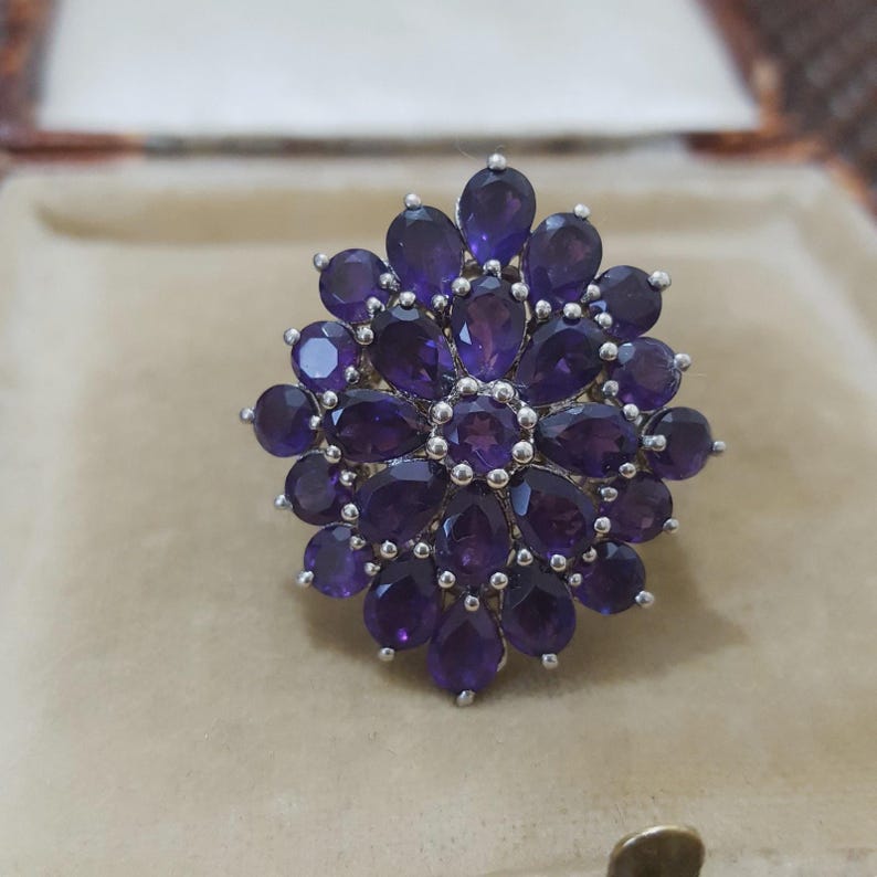 K&ouml;nnte beinhalten: Ein silberner Ring mit zahlreichen tiefvioletten Amethyst-Edelsteinen. Die Edelsteine sind in einem floralen Muster angeordnet, mit einem zentralen Cluster, der von tropfenf&ouml;rmigen Steinen umgeben ist. Der Ring ist vor einem neutralen Hintergrund platziert.