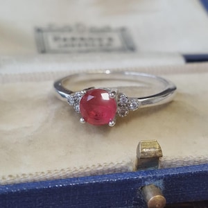 May include: A silver-toned ring featuring a round, faceted, deep pink gemstone set in the centre. The band is accented on both sides of the central stone by three small, clear, round-cut stones arranged in a cluster, set within a metallic frame.