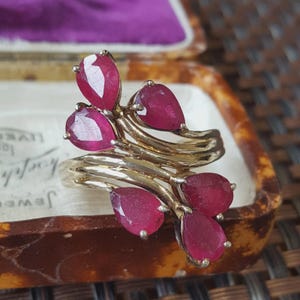 May include: A gold ring featuring five pear-shaped, deep red rubies arranged in a cluster. The band consists of multiple polished gold strands that curve and intertwine to support the faceted gemstones, creating a layered, organic appearance.