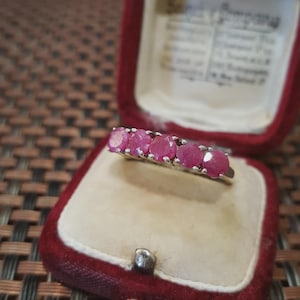 May include: A silver ring with five oval-cut, vibrant pink gemstones. The ring is displayed inside a red velvet jewelry box with a cream-colored interior, showcasing the ring's sparkle and color.