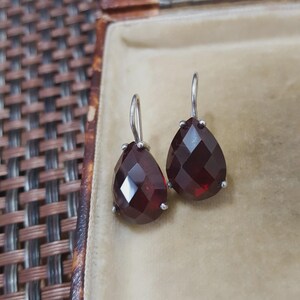 May include: A pair of silver drop earrings featuring faceted, teardrop-shaped, deep red gemstones. The earrings are presented inside a vintage, tan-coloured jewellery box with a woven brown exterior.