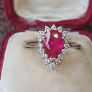May include: A silver-toned ring featuring a central pear-shaped red gemstone surrounded by a halo of small, clear, round-cut stones. The ring has a polished metal band and is presented in a deep red, velvet-lined jewellery box.