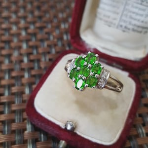 May include: A silver ring with a cluster of oval-cut, bright green gemstones and small clear stones. The ring is displayed in a red velvet ring box with a cream-colored interior.
