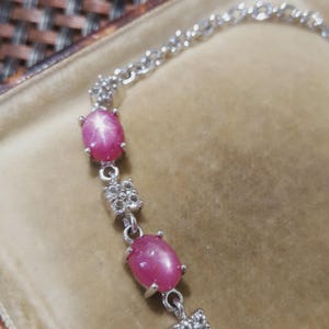 May include: A silver bracelet featuring oval, pink gemstones with a star-like effect. The bracelet has a delicate chain and decorative links with small, clear stones. The gemstones are set in a silver metal.