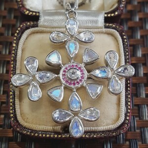May include: A silver cross pendant featuring multiple pear-shaped moonstones with iridescent blue flashes. The center of the cross is accented by a circular arrangement of small, vibrant pink gemstones surrounding a single clear, round-cut stone.