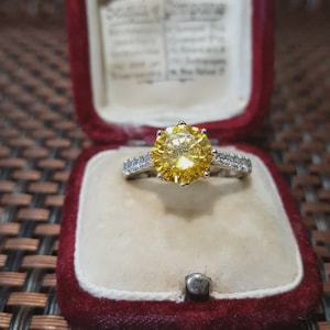 Può includere: Anello d'argento con una grande gemma gialla rotonda. La fascia è adornata da pietre più piccole e trasparenti. L'anello è presentato in un cofanetto di velluto rosso con interno color crema.