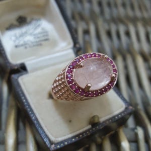 May include: Rose gold ring featuring a large oval, pale pink gemstone, encircled by small red gemstones. The ring has a textured band and is displayed in a vintage jewellery box.