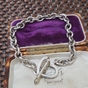 May include: A silver chain bracelet with a heart-shaped clasp. The bracelet is made of thick, interlocking links. The clasp is a heart with a bar that slides through to secure the bracelet. The bracelet is displayed in a vintage jewelry box.