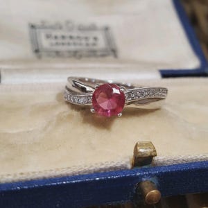 Könnte beinhalten: Ein silberfarbener Ring mit einem runden, facettierten, leuchtend rosa Mittelstein, der von vier Krappen gehalten wird. Die Ringschiene ist geteilt und mit einer Reihe kleiner, klarer, funkelnder Steine an den Seiten besetzt, was für ein poliertes Aussehen sorgt.