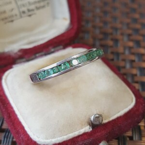Peut inclure: Une bague en métal argenté dotée d'un sertissage rail avec une rangée de petites pierres précieuses rondes d'un vert éclatant. La bague est placée à l'intérieur d'une petite boîte à bijoux en velours rouge avec un coussin intérieur crème.