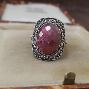 Op de afbeelding: Een zilverkleurige ring met een grote, facetgeslepen, ovaalvormige, dieprode edelsteen. De edelsteen is omgeven door een rand van kleine, ronde, zilverkleurige stenen. De ring is gezet in een rechthoekig frame met decoratieve details.
