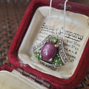 May include: A silver pendant necklace featuring a central oval, purplish-red gemstone, encircled by smaller green gemstones and clear accent stones. The necklace is presented in a vintage red jewellery box.