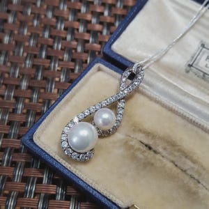 May include: A silver pendant necklace featuring two pearls and a diamond-set design. The necklace is presented in a blue jewellery box with a light beige interior. The pearls are white and the diamonds sparkle.