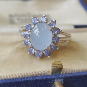 May include: A silver ring featuring a large, oval, light blue cabochon gemstone at the centre, surrounded by a halo of ten smaller, round, periwinkle-coloured faceted gemstones. The ring has a polished silver band and a multi-pronged setting.
