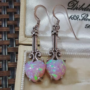 Può includere: Un paio di orecchini pendenti in argento con pietre di opale rosa a forma di cuore e macchie verdi iridescenti. Gli orecchini presentano un perno in metallo decorativo con volute e un gancio curvo per l'orecchio.