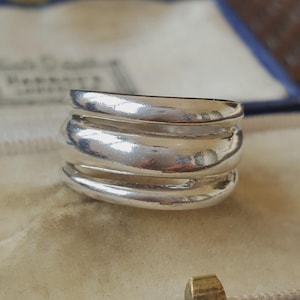 May include: A polished silver ring featuring a multi-band design composed of three distinct, smooth, rounded horizontal bands that are connected to form a single cohesive piece of jewellery with a reflective, metallic finish and open spaces.