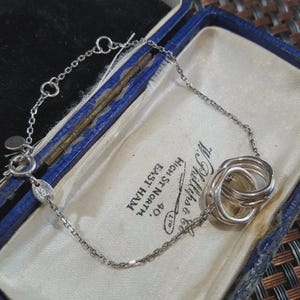 May include: A silver chain bracelet featuring three interlocking silver rings as a central charm. The bracelet includes a delicate link chain, a circular clasp, and a small oval tag with engraved text. The jewellery is presented in a blue box.
