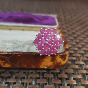 Może przedstawiać: Srebrny pierścionek z klastrem małych, okrągłych, jasnoróżowych kamieni szlachetnych. Kamienie szlachetne ułożone są we wzór kwiatowy. Pierścionek umieszczony jest w zabytkowym pudełku na biżuterię z fioletowym wnętrzem.