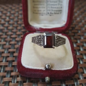 Peut inclure: Bague en argent avec une pierre précieuse rectangulaire rouge foncé, présentée dans un écrin à bijoux en velours rouge vintage. La monture de la bague présente un motif texturé de chaque côté de la pierre, pour un look classique et élégant.