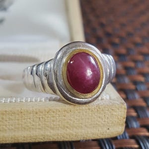 Può includere: Un anello in argento con una pietra preziosa rosso scuro a taglio cabochon ovale al centro. La pietra è incastonata in una sottile lunetta color oro, circondata da una cornice in argento più ampia con un design a fascia scanalata e testurizzata.