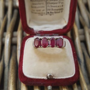 May include: A gold ring with four oval-cut, deep red gemstones. The ring is displayed inside a vintage red velvet jewelry box with a cream-colored interior. The box is open, revealing the ring against the soft fabric.