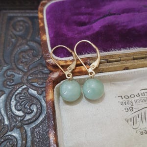 May include: Gold-coloured leverback earrings featuring pale green jade spheres. The earrings are presented in a vintage jewellery box with a purple velvet interior.