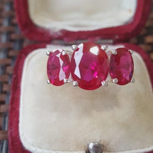 May include: A silver ring featuring three oval-cut, vibrant red gemstones arranged in a row. The centre stone is larger than the two flanking stones, and each gem is held in place by four silver prongs, creating a classic three-stone design.