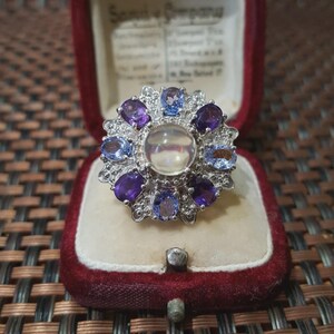 May include: A silver ring featuring a central iridescent stone, encircled by alternating purple and blue gemstones. The ring has a decorative, floral design and is presented in a red velvet jewellery box.