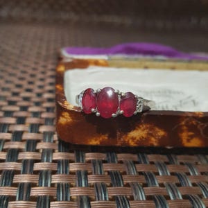 Op de afbeelding: Een zilveren ring met drie ovale, dieprode edelstenen. De ring heeft kleine, heldere accentstenen. De ring wordt getoond in een vintage sieradendoosje met een bruin en tan schildpadpatroon.