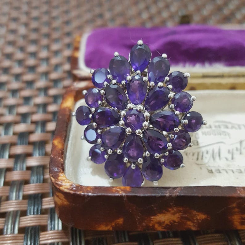 K&ouml;nnte beinhalten: Silberfarbene Brosche mit floralem Design, besetzt mit zahlreichen tiefvioletten Amethyst-Edelsteinen. Die Brosche wird in einer Vintage-Schmuckschatulle mit violettem Samt pr&auml;sentiert.