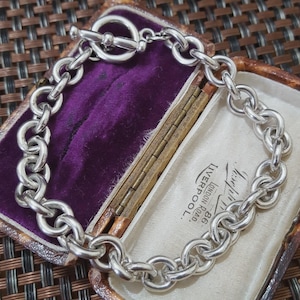Op de afbeelding: Een zilveren kettingarmband met een tuimelsluiting, gepresenteerd in een vintage sieradendoosje met een paars fluwelen interieur. De armband heeft grote, afgeronde schakels.