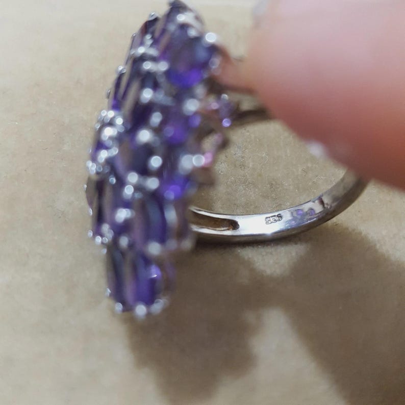 K&ouml;nnte beinhalten: Ein silberner Ring mit einem Cluster aus oval geschliffenen, lila Edelsteinen. Der Ring hat ein glattes, poliertes Band mit einem innen sichtbaren Stempel. Die Edelsteine sind in einem blumen- oder blattartigen Muster angeordnet.