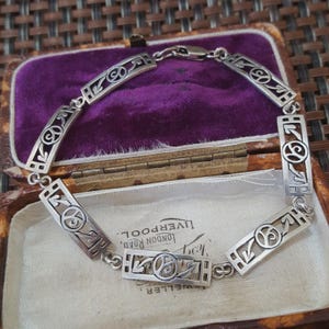 May include: A silver bracelet with a series of rectangular and curved links, each featuring an openwork design. The bracelet is displayed inside a vintage jewelry box with a purple velvet interior.