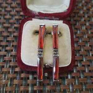 May include: Silver earrings featuring rectangular, deep red gemstones. The earrings are presented in a vintage, burgundy velvet jewellery box. The gemstones are set in a linear design, offering a sleek and elegant appearance.
