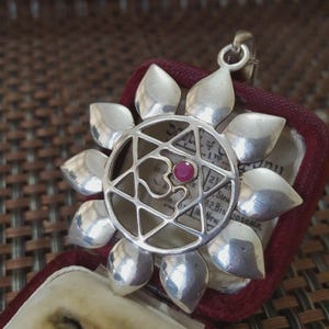 Op de afbeelding: Zilveren hanger met een bloemachtig ontwerp. De hanger heeft een centraal geometrisch patroon met een kleine, ronde, rode edelsteen. De bloemblaadjes zijn glad en afgerond. De hanger wordt gepresenteerd in een bordeauxrode fluwelen doos.