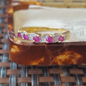 May include: A gold band ring featuring four round, vibrant pink rubies set in a row, alternating with small, clear, sparkling accent stones. The setting is crafted with a textured, silver-toned metal finish surrounding each of the primary stones.