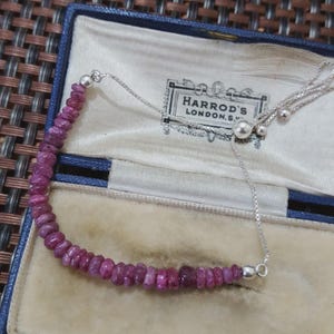 May include: A delicate necklace featuring a strand of small, irregular, deep pink gemstones. The necklace has a silver chain and a sliding bead closure. The necklace is displayed inside a blue jewelry box with the Harrods London logo.
